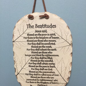 May include: Oval-shaped, off-white plaque with a brown leather cord for hanging. The plaque features the text "The Beatitudes" and a passage from Matthew 5, with leaf vein designs. A small dragonfly is at the bottom.