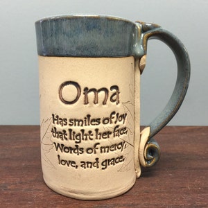 May include: A ceramic mug with a blue rim and handle, featuring the word "Oma" and an inscription about joy, mercy, love, and grace. The mug has a beige body with leaf-like designs.