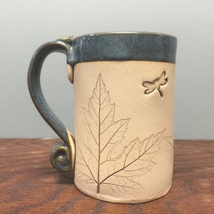 May include: Ceramic mug with a cream-colored body and a teal blue rim and handle. The mug features an embossed leaf and dragonfly design. The handle has a decorative swirl at the base.