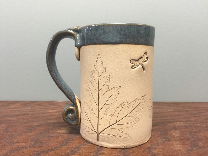 May include: Ceramic mug with a cream-colored body and a teal blue rim and handle. The mug features an embossed leaf and dragonfly design. The handle has a decorative swirl at the base.