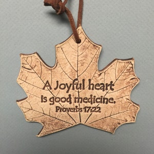 May include: A leaf-shaped decorative plaque with the text "A Joyful heart is good medicine. Proverbs 17:22." The plaque is light brown with dark brown veining and a brown leather cord for hanging.