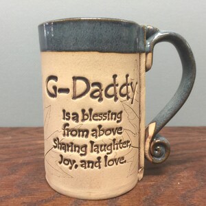 May include: A handcrafted ceramic mug with a beige body and a blue rim and handle. The mug features the inscription "G-Daddy is a blessing from above Sharing laughter, Joy, and love." Decorative leaf patterns are etched on the mug.