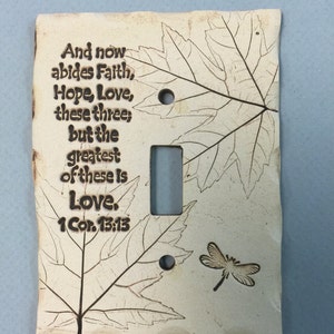 May include: A cream-colored ceramic light switch cover with engraved leaf designs and the text "And now abides Faith, Hope, Love, these three; but the greatest of these is Love. 1 Cor. 13:13". A dragonfly is also engraved.