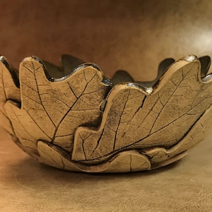 May include: A handcrafted ceramic bowl shaped like a leaf, with intricate vein details. The bowl has a light brown color with darker accents, and a scalloped edge. This decorative bowl is ideal for serving or display.