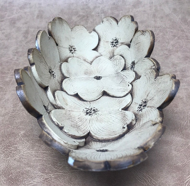 May include: A decorative bowl shaped like overlapping flowers. The bowl is painted in a cream color with brown edges, and each flower has a detailed center. The bowl is designed to hold small items and adds a touch of nature-inspired decor.