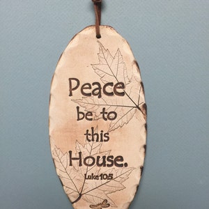 May include: Oval-shaped ceramic plaque with the words "Peace be to this House" and "Luke 10:5" in dark brown lettering. The plaque features leaf and dragonfly designs. A leather cord is attached for hanging.