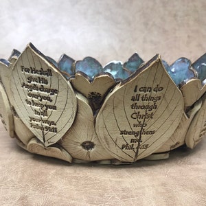 May include: A ceramic bowl with a leaf and flower design. The bowl is beige with blue accents and features engraved text from the Bible. The text includes "For He shall give His Angels charge over you..." and "I can do all things through Christ..."