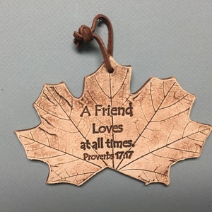 May include: A leaf-shaped ornament with the text "A Friend Loves at all times. Proverbs 17:17" in a brown, textured finish. It has a leather loop for hanging. The ornament is on a light blue background.