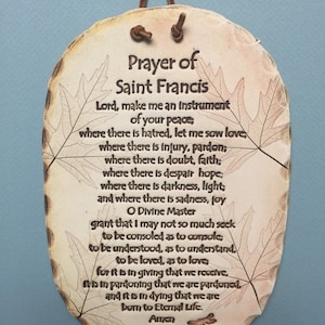 May include: Oval-shaped plaque with the "Prayer of Saint Francis" printed in black. The plaque has a light brown, wood-like appearance with leaf designs and a brown leather cord for hanging.