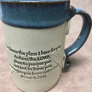 May include: A handmade ceramic mug with a blue rim and handle, and a cream-colored body. The mug features an inscription of Jeremiah 29:11. The mug has a unique, textured surface and a curved handle.