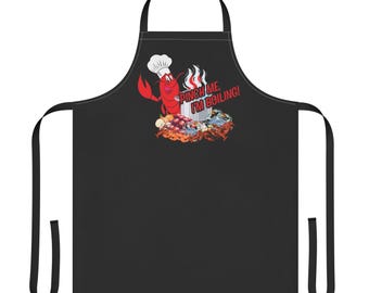 May include: A black apron featuring a graphic of a red lobster wearing a white chef hat next to a steaming pot. The text PINCH ME, I'M BOILING! is printed in red above a pile of various seafood including crawfish, crabs, and onions.