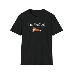 Op de afbeelding: Zwart T-shirt met witte tekst die "I'm Sheltied" zegt en een illustratie van een bruine en witte Shetland Sheepdog.