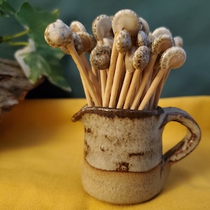 May include: A ceramic pitcher with a brown glaze and a speckled pattern holds a bunch of wooden skewers topped with white ceramic mushroom-shaped heads.