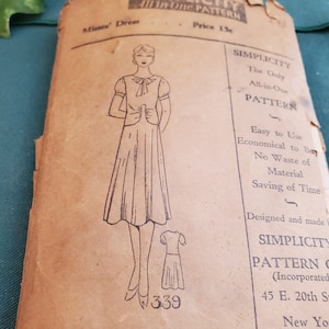 May include: Vintage Simplicity pattern for a dress, pattern number 339. The pattern is for a simple, elegant dress with a bow at the neckline. The pattern is printed on a brown paper background.