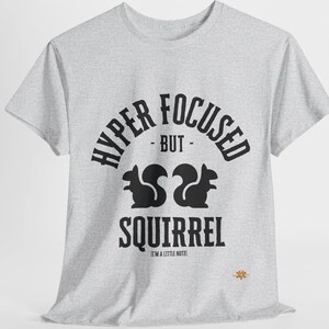 Op de afbeelding: Lichtgrijs t-shirt met zwarte tekst "HYPER FOCUSED - BUT - SQUIRREL" en twee silhouetten van eekhoorns. De tekst "I'M A LITTLE NUTZ!" staat onder het woord "SQUIRREL".