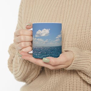 May include: White ceramic mug featuring a vibrant image of a blue ocean under a bright blue sky with fluffy white clouds. The mug is held by a person wearing a beige sweater.