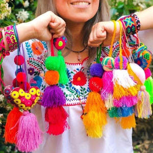 May include: A collection of colorful handmade tassels and pom-poms in various sizes and vibrant colors, including pink, orange, green, and purple. The tassels are suspended from cords and are being held up by a person. The person is wearing a white embroidered top and bracelets.