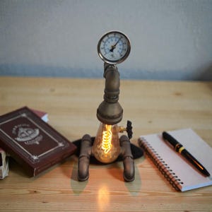 Puede incluir: Una lámpara de escritorio de estilo steampunk con un manómetro y una bombilla expuesta. La lámpara está construida con tuberías y accesorios de latón, con un filamento cálido y brillante. Un cuaderno, un bolígrafo y un libro también están sobre la superficie de madera.
