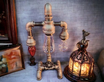 May include: A steampunk-style lamp made from metal pipes and fittings, featuring three Edison-style light bulbs. The lamp has a unique, industrial design with a valve detail at the base. A red glass bulb and a decorative lamp are also visible.