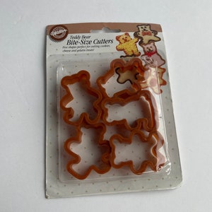 Peut inclure: Un paquet de cinq emporte-pièces en plastique brun en forme d'ours en peluche. L'emballage a un fond blanc avec des pois noirs et le texte "Teddy Bear Bite-Size Cutters" en brun. Le texte "Five shapes perfect for cutting cookies, cheese and gelatin treats!" est également imprimé sur l'emballage.