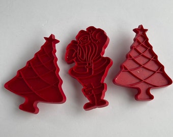 May include: Three red plastic Christmas cookie cutters. One cutter is shaped like Santa Claus, and the other two are shaped like Christmas trees.