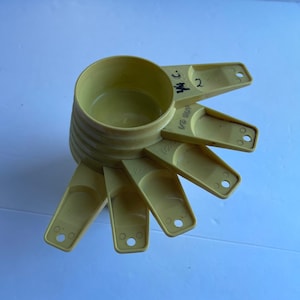May include: A set of six yellow plastic measuring cups. The cups are nested together and have handles. The largest cup is labelled "1/2 cup" and the smallest cup is labelled "1/4 cup".