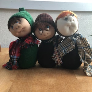 May include: Three handmade plush toys with different colored hats and scarves. The toys are made of fabric and have a round, slightly misshapen head. The first toy is wearing a green hat and a red and green plaid scarf. The second toy is wearing a brown hat and a black and white plaid scarf. The third toy is wearing a red hat and a blue and white plaid scarf.