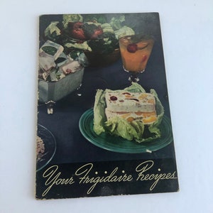 May include: A vintage cookbook with a green and white dessert on a green plate, a glass of orange juice with a cherry, and a bowl of ice cubes. The cookbook cover reads "Your Frigidaire Recipes".