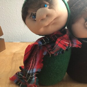 May include: Two handmade plush dolls with green and brown knit hats, one with a red and black plaid scarf. The dolls have painted eyes and a stitched facial expression.