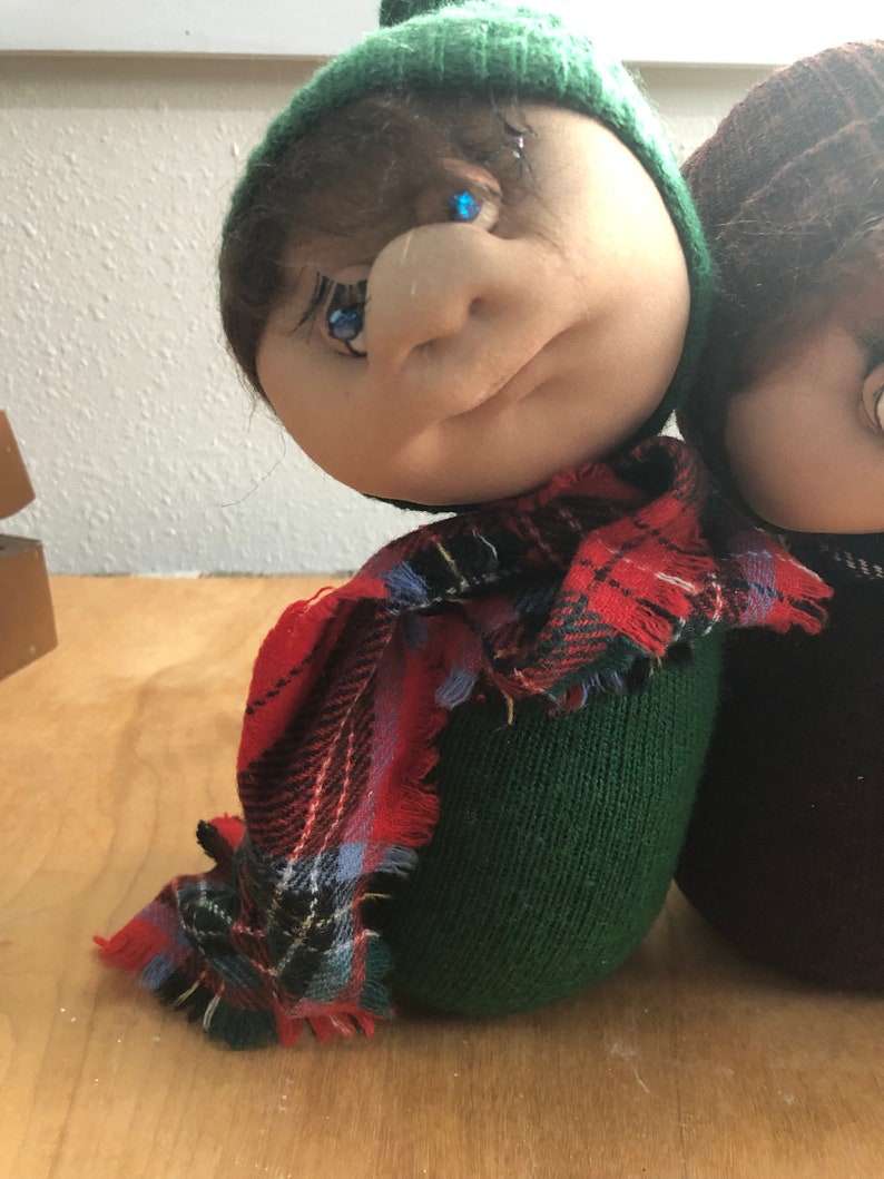 May include: Two handmade plush dolls with green and brown knit hats, one with a red and black plaid scarf. The dolls have painted eyes and a stitched facial expression.
