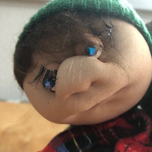 May include: A close-up of a doll's face with brown hair, a green knit hat, and blue eyes. The doll is wearing a red and black plaid shirt.