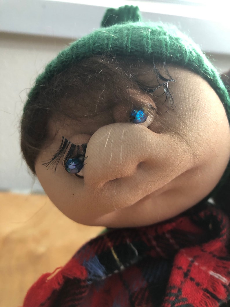 May include: A close-up of a doll's face with brown hair, a green knit hat, and blue eyes. The doll is wearing a red and black plaid shirt.