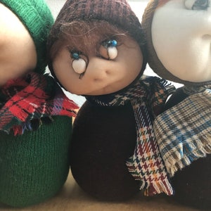 May include: Three handmade plush dolls wearing hats and scarves. The dolls are brown, green, and white with blue eyes.