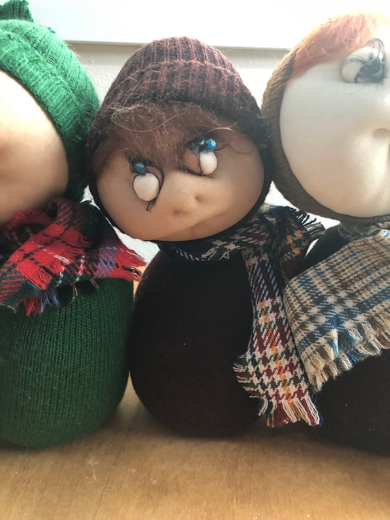May include: Three handmade plush dolls wearing hats and scarves. The dolls are brown, green, and white with blue eyes.