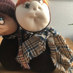 May include: Two handmade fabric dolls with brown and white knit hats and scarves. The dolls have large eyes and are wearing plaid scarves.