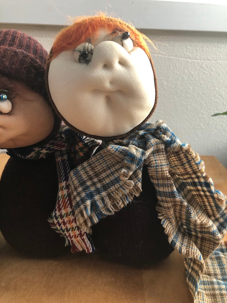 May include: Two handmade fabric dolls with brown and white knit hats and scarves. The dolls have large eyes and are wearing plaid scarves.