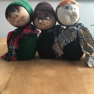 May include: Three handmade fabric dolls with different colored hats and scarves. The dolls have painted eyes and are wearing plaid scarves.