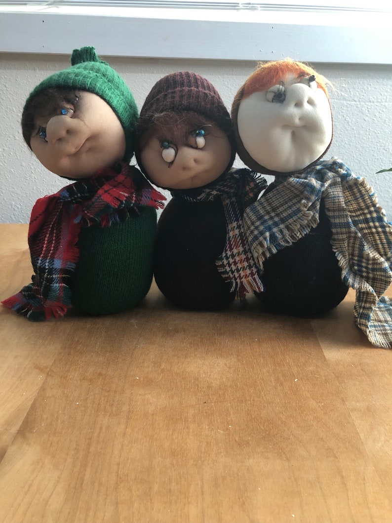 May include: Three handmade fabric dolls with different colored hats and scarves. The dolls have painted eyes and are wearing plaid scarves.