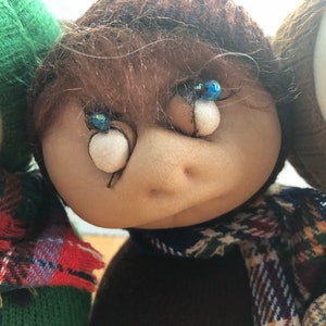 May include: A brown-haired doll with blue eyes and a plaid scarf. The doll is wearing a brown sweater and a brown hat.
