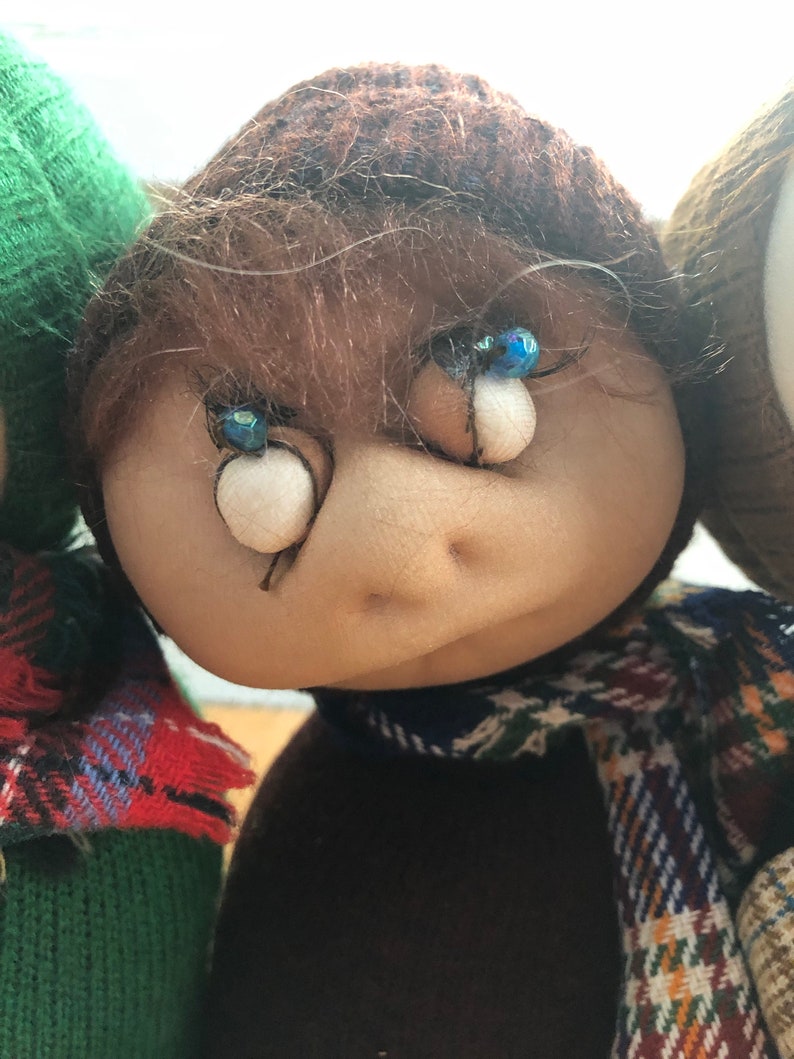 May include: A brown-haired doll with blue eyes and a plaid scarf. The doll is wearing a brown sweater and a brown hat.