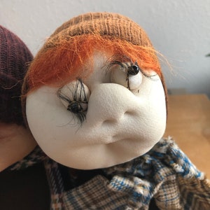 May include: A handmade doll with a brown knit hat, orange hair, and a plaid scarf. The doll has a white face with black eyelashes and brown eyes.