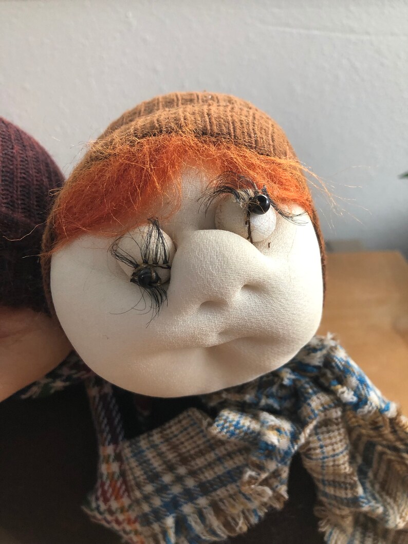 May include: A handmade doll with a brown knit hat, orange hair, and a plaid scarf. The doll has a white face with black eyelashes and brown eyes.