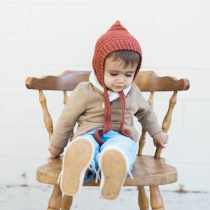 May include: A toddler wearing a brown knitted hat with a pointed top and blue jeans sits on a wooden chair.