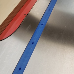May include: A close-up shot of a blue, rectangular piece of material with several evenly spaced holes. The piece is positioned diagonally across a metallic surface. A red and brown object is visible in the upper left corner.