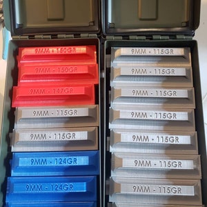 May include: Two open olive green plastic ammunition storage boxes containing multiple 3D-printed plastic ammunition organizers. The organizers are labeled with text including 9MM - 150GR, 9MM - 147GR, 9MM - 124GR, and 9MM - 115GR in red, blue, and grey colors.