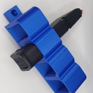 Może przedstawiać: Jasnoniebieski uchwyt na magazynki wydrukowany w 3D. Uchwyt jest przeznaczony do przechowywania dwóch magazynków, z jednym czarnym magazynkiem włożonym. Uchwyt ma mały, oddzielny element z otworem. Uchwyt jest wykonany z tworzywa sztucznego.