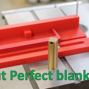 Peut inclure: Un gabarit de menuiserie rouge avec une ébauche en laiton et en bois, positionné sur une scie à table. Le texte "Cut Perfect blanks!" est affiché en vert en bas de l'image. Le gabarit semble conçu pour une coupe précise.
