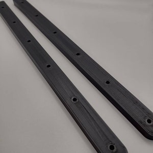 May include: Two long, dark gray, rectangular pieces with rounded ends and multiple evenly spaced holes. The pieces have a ridged texture and appear to be made of plastic or a similar material. They are positioned diagonally against a light gray background.