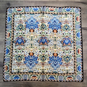May include: A square silk scarf featuring a repeating botanical pattern with large blue flowers, green foliage, and orange accents on a white background. The scarf has a decorative border with a black edge and the word LIBERTY printed in the corner.