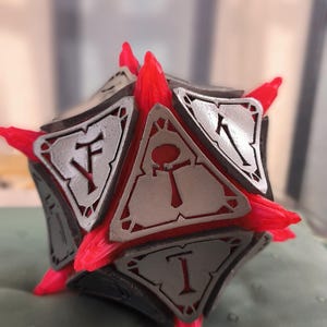 Può includere: Un oggetto geometrico sfaccettato con una base grigio scuro, pannelli argentati e sporgenze rosse e appuntite. Ogni pannello presenta un simbolo. L'oggetto è probabilmente un oggetto decorativo o un'opera d'arte unica.